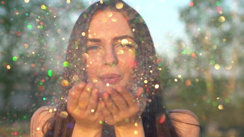 Beautiful woman blowing confetti in slow motion on nature beach. Caucasian happy teenage model girl blowing gold glitter off hands in summer park at sunset. Simmertime Outside Beauty happiness summer
