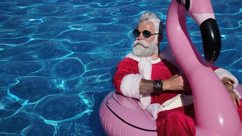 Handsome Santa floating in flamingo inflatable ring into the swimming pool with cocktail glass in hand, chilling and enjoying tropical New Year. Pleasure and happiness concept. Inspiration background