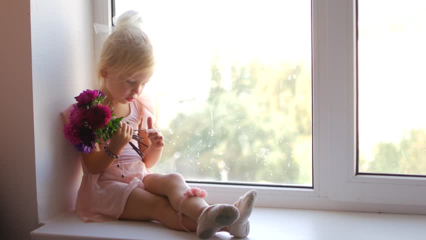 The young ballerina sits on the window and holds flowers in her hands