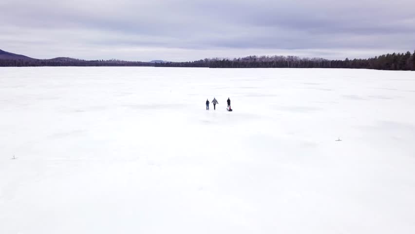 Get an aerial view of Ice Fishing on Fitzgerald Pond, Maine. We pass three people pulling a sled.