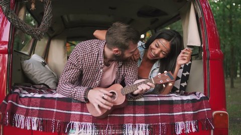 Happy young mixed race couple playing instruments and having some fun in retro hippie minibus in forest, slow motion, close up