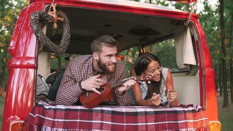 Happy young mixed race couple playing instruments and having some fun in retro hippie minibus in forest, slow motion