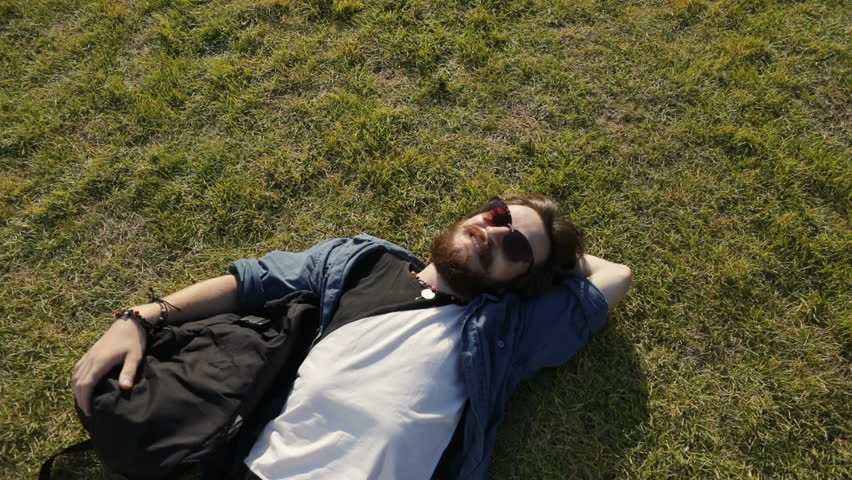 Relaxed, joyful biker resting on green grass, lying near by bike peacefully after hard working day, outdoor shot in urban, recreation area