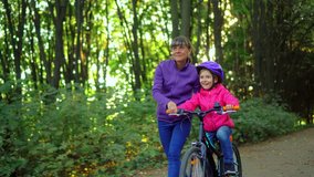 Mother teaching daughter to ride bike at urban park. Child girl learning biking with the mom  help.Family and childhood concept.Sunny autumn day.Side gimbal follow 4k video