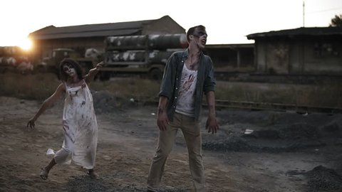 Creepy two zombies in bloody clothes walking through the ruined city during the zombie apocalypse. Abandoned place with trucks on the background