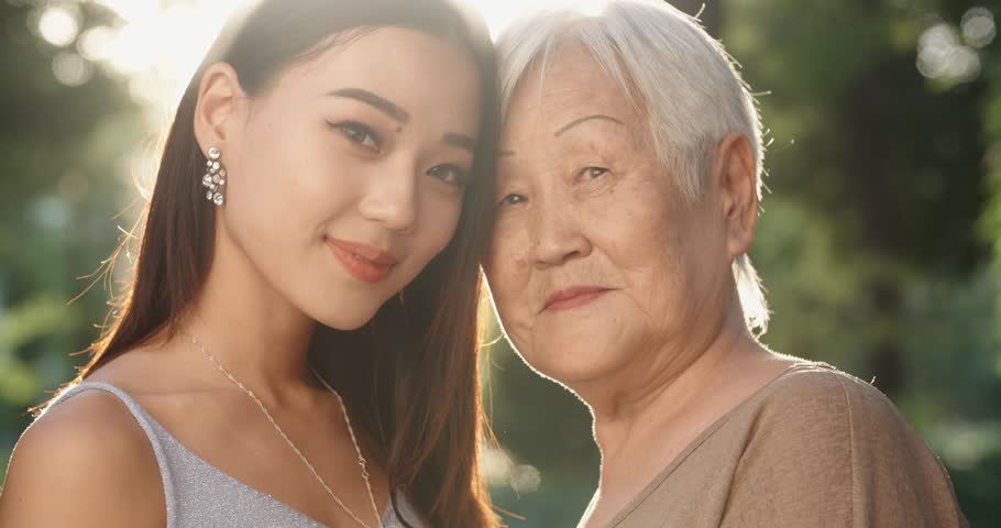 Happy asian grandmother and her teen granddaughter standing in park, hugging each other and smiling - portrait shot 4k