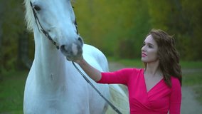 Young romantic girl in red dress walking with white horse against the sun. Woman Rider with her stallion in autumn forest at sunset. Slow motion. Sun lens flare