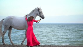 Young romantic girl in red dress walking with white horse against the sun on the background of the sea. Woman Rider with her stallion in beach. Slow motion. Sun lens flare