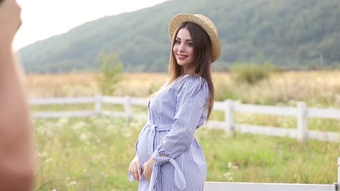 Pregnant woman in blue dress and knitted hat poses to camera