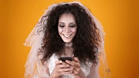 Happy dead bride on halloween wearing wedding dress and makeup using smartphone over orange background