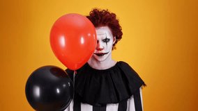 Cheerful clown with halloween makeup having fun with balloons and looking at the camera over orange background
