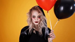 Happy woman wizard wearing black costume and halloween makeup posing with balloons and looking at the camera over orange background