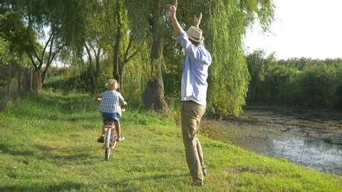 little boy learning to ride a bicycle with help from her dad on background outdoor nature and small river