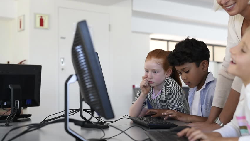 Teacher helping multiethnic children at computer terminal in primary school. Smiling school children looking at computer screen with teacher in library. Computer class at elementary school. Royalty-Free Stock Footage #1017377782