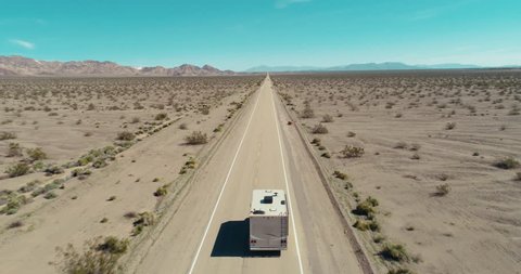 AERIAL - A drone follows a brown camper van from the back in the middle of the Mojave Desert and slowly gets closer to the van on route 66 on a clear blue day.