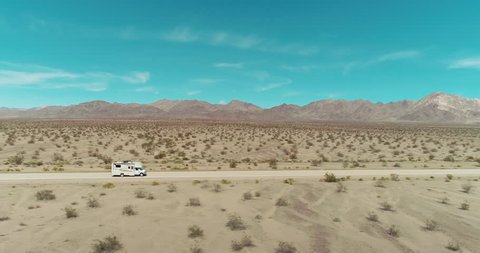AERIAL - A brown camper van in the distance drives from left to right through the image while being passed by a small black car in the middle of the Mojave desert on a clear and blue day