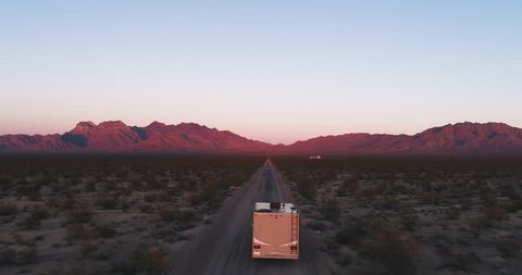 AERIAL - The camera follows an RV that is being lit by the last bit of sun during sunset while driving towards some mountains in the desert