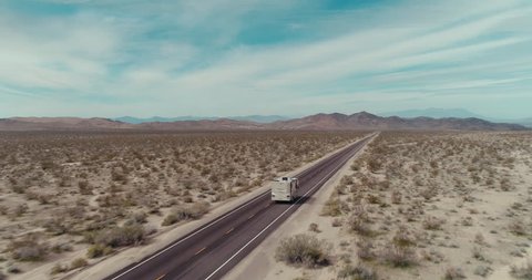 AERIAL - A drone follows a brown RV in the Mojave desert on a sunny day with mountains in the back and tumbleweed next to the road