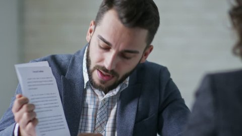 Young bearded businessman giving two paged document with terms and conditions printed on it to unrecognizable female client seen from her back