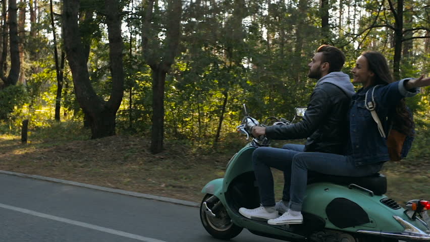 Young, happy couple riding on the scooter in the forest. Happy woman having fun and spreading hands. View from aside.
