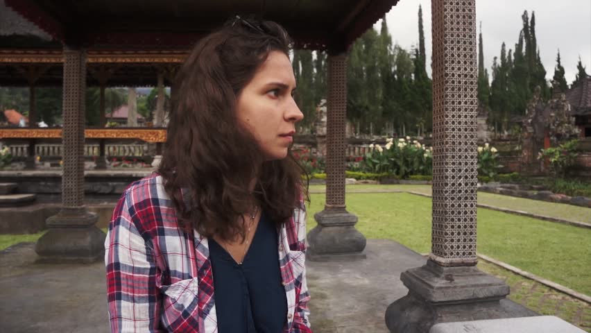 Attractive young woman with dark curly hair wearing colored short is sitting in wooden and stone carved arbor in balinese park on cloudy summer day.