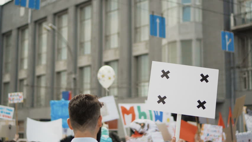 Angry adult young man with a banner in hands go and shouts loudspeaker rally. Aggressive protester on strike. Revolution in city during day. Crowd people in the background on street. 20s caucasion boy