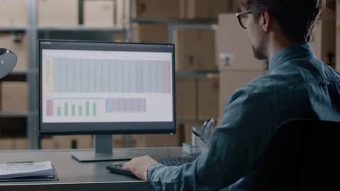Warehouse Inventory Manager Works with a Spreadsheet on a Personal Computer while Sitting at His Desk. In the Background Shelves Full of Cardboard Box Packages Ready For Shipping.Shot on RED EPIC-W 8K
