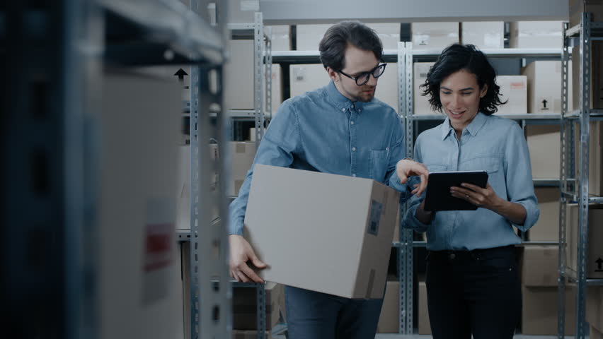 Female Inventory Manager Shows Digital Tablet Information to a Worker Holding Cardboard Box, They Talk and Do Work. In the Background Stock of Parcels with Products Ready for Shipment.  Royalty-Free Stock Footage #1017422854