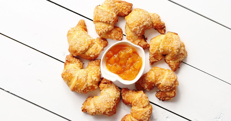 From above shot of small delicious croissants lying around bowl of sweet jam on surface of white wooden tabletop