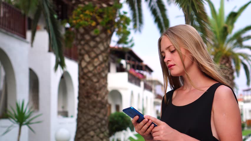 Dreamy busy relaxed young girl looking at smart phone device screen reading in middle of palm tree tropical resort alley