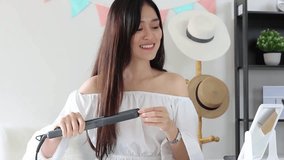 Young Beautiful Asian beauty blogger woman in white room straightening long hair with iron and smiling.