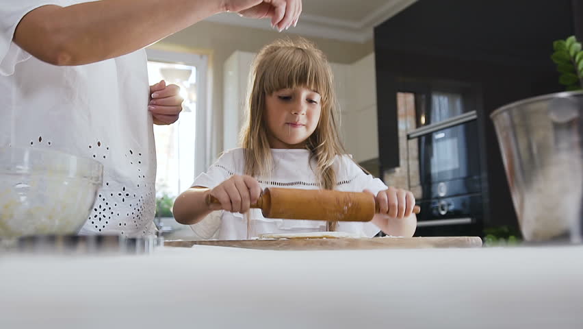 Good looking mother teaching her young little daughter to cook and kneading a dough for cookies on the table in the kitchen at home. Young woman and small girl preparing pasta or pizza together using Royalty-Free Stock Footage #1017464338