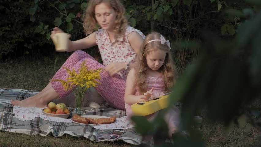 A happy mother and daughter are playing on the ukulele. Family in a city park on a picnic on a warm evening at sunset. 4k