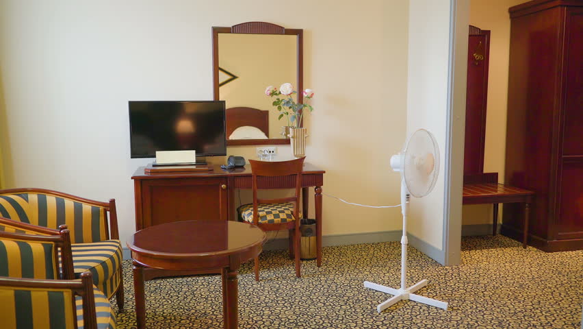 The view inside the bedroom of the hotel with the stand fan moving left and right and the dresser with the mirror and the chair on the side