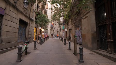 BARCELONA, SPAIN - AUGUST 13, 2018: Walking up along nice Carrer d'Ataulf, old street at Gothic Quarter of Barcelona, first person view. Small trees and bollards at sides, closed doors of shops