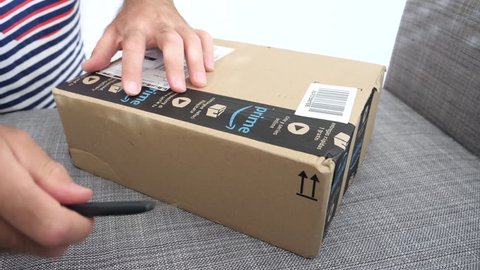 MUNICH, GERMANY - CIRCA 2018: Man unboxing on the sofa the newly arrived package parcel from Amazon Prime - curiosity opening the box with cutter