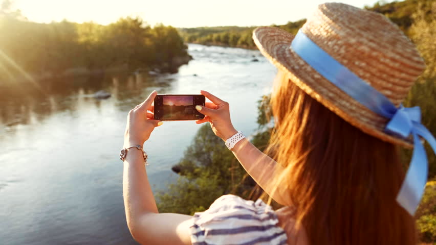 Woman traveling, making photo of beautiful river and mountains nature landscape on smart phone. Traveler girl using smartphone walking outdoor in park, taking mobile picture of sunset forest.