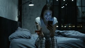 Young beautiful female having late night video call, using her smartphone, sitting on bed, drinking wine. Long distance relationship with boyfriend concept