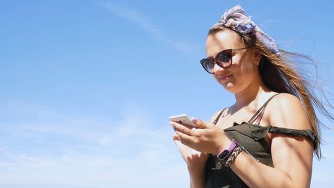 Happy woman in glasses uses a smartphone, on the beach in windy weather, in slow motion