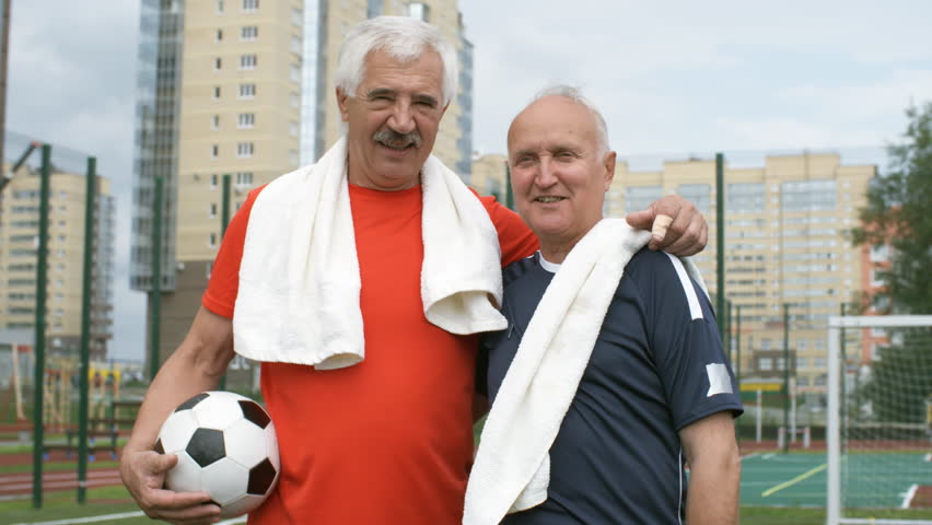 Medium shot of two optimistic elderly male friends with towels on shoulders hugging and talking to each other while standing on football field after game, one of them holding ball