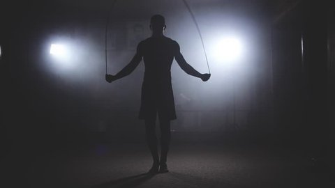 Boxer does some jump rope exercises in dark room under light. Fighter training in smoky studio in slow motion. Silhouette on dark background