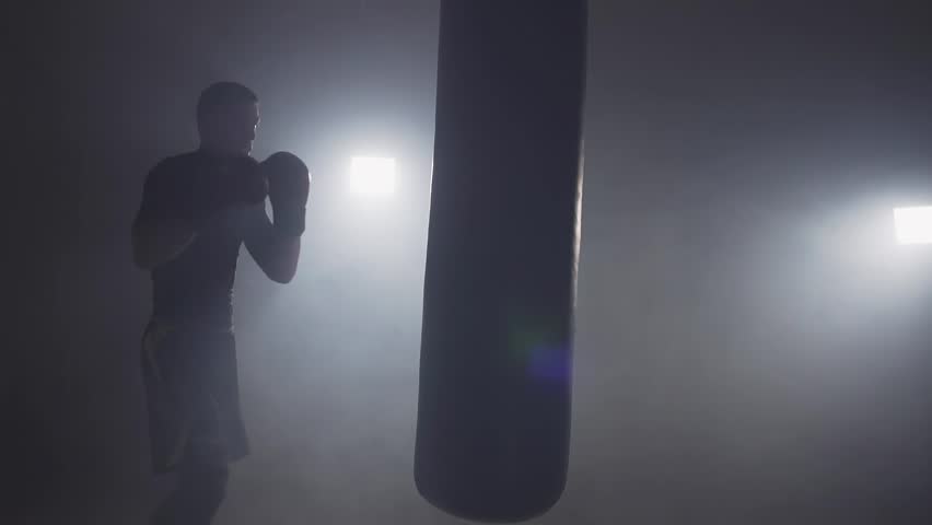 Alone boxer hits punching bag in dark gym in slow motion. Young man training indoors. Strong athlete in gym. Sport concept. Medium shot. Sportsman boxing in smoky studio Royalty-Free Stock Footage #1017517615