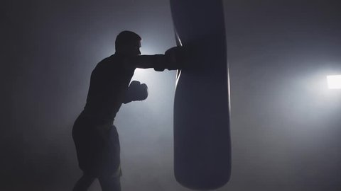 Alone boxer hits punching bag in dark gym in slow motion. Young man training indoors. Strong athlete in gym. Sport concept. Medium shot. Sportsman boxing in smoky studio