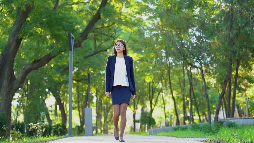 Young beautiful happy business woman in formal wear and glasses walking in the green park alone. Girl keeps way to work or university