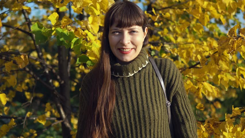 A young woman dressed in a green knitted sweater shows thumbs up against a background of yellowed trees.