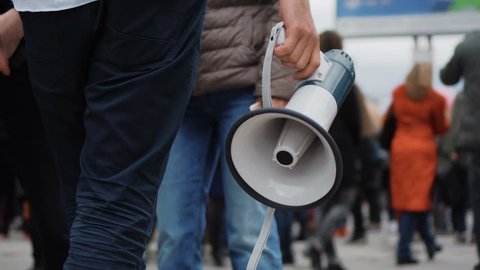 1 Man holds a megaphone in his hands, walking down street through many people on strike. Close up of one guy with loudspeaker in crowd at a demonstration 4k. Young adult boy on revolution.