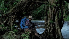 Adventurers Asian men. sitting enjoying and looking beautiful waterfall. Travel Lifestyle concept vacations into the wild wearing cozy shirt,hat with backpack. video 4k  Slow motion