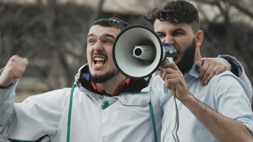 Two guys on strike shouting into a megaphone close-up. People are yelling on the street. Angry football fans in casual wear. Cool plan. Serious boys slow motion demonstration. Political strike out.