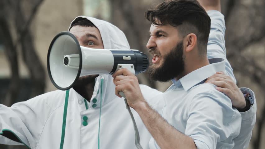 Two guys on strike shouting into a megaphone close-up. People are yelling on the street. Angry football fans in casual wear. Cool plan. Serious boys at a slow motion demonstration. Royalty-Free Stock Footage #1017522505