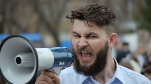A desperate man goes and demands a megaphone on strike. European guy with a beard is yelling into the loudspeaker at a rally. People are unhappy with the government. Revolution on the street in city.
